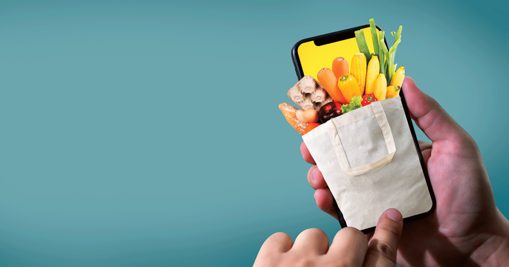 Pessoa utilizando um aplicativo personalizado de supermercado no celular, com sacola virtual de compras repleta de alimentos frescos