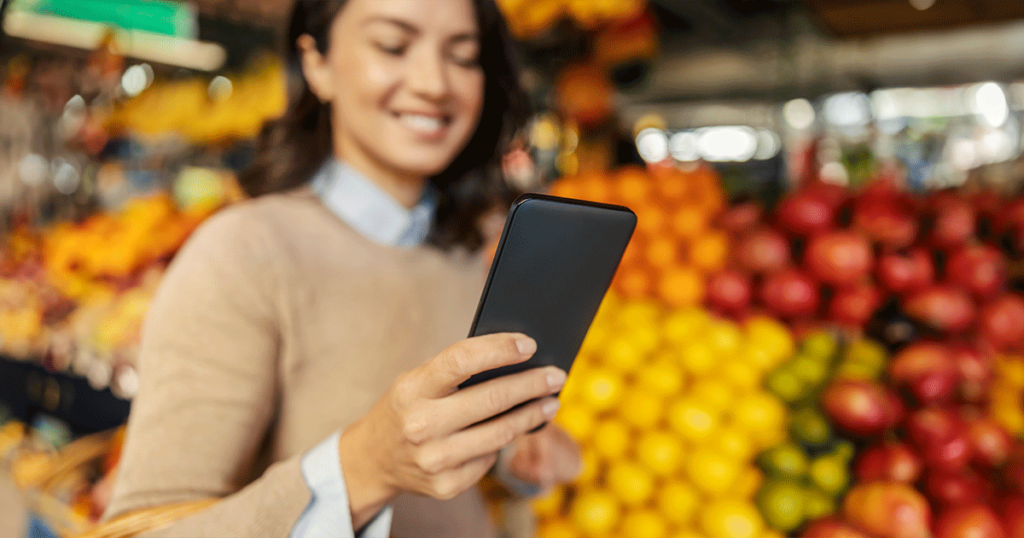 Mulher dentro de um supermercado conferindo no celular o aplicativo de supermercado para verificar ofertas e produtos disponíveis.