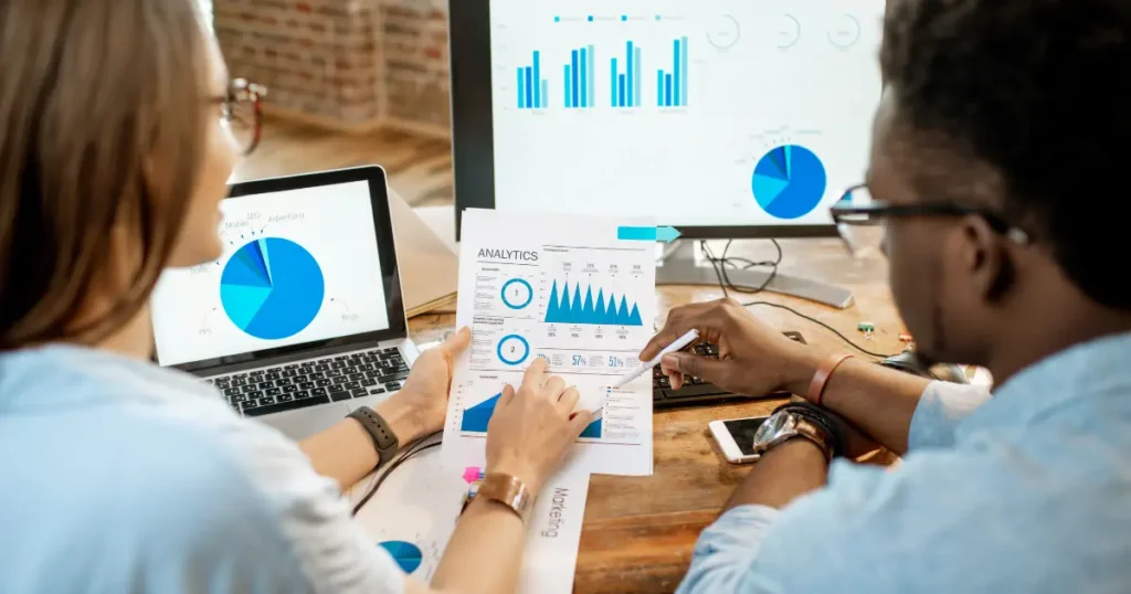 Dois profissionais analisando gráficos e relatórios detalhados em computadores e documentos, destacando a importância da análise de dados para lojas autônomas.